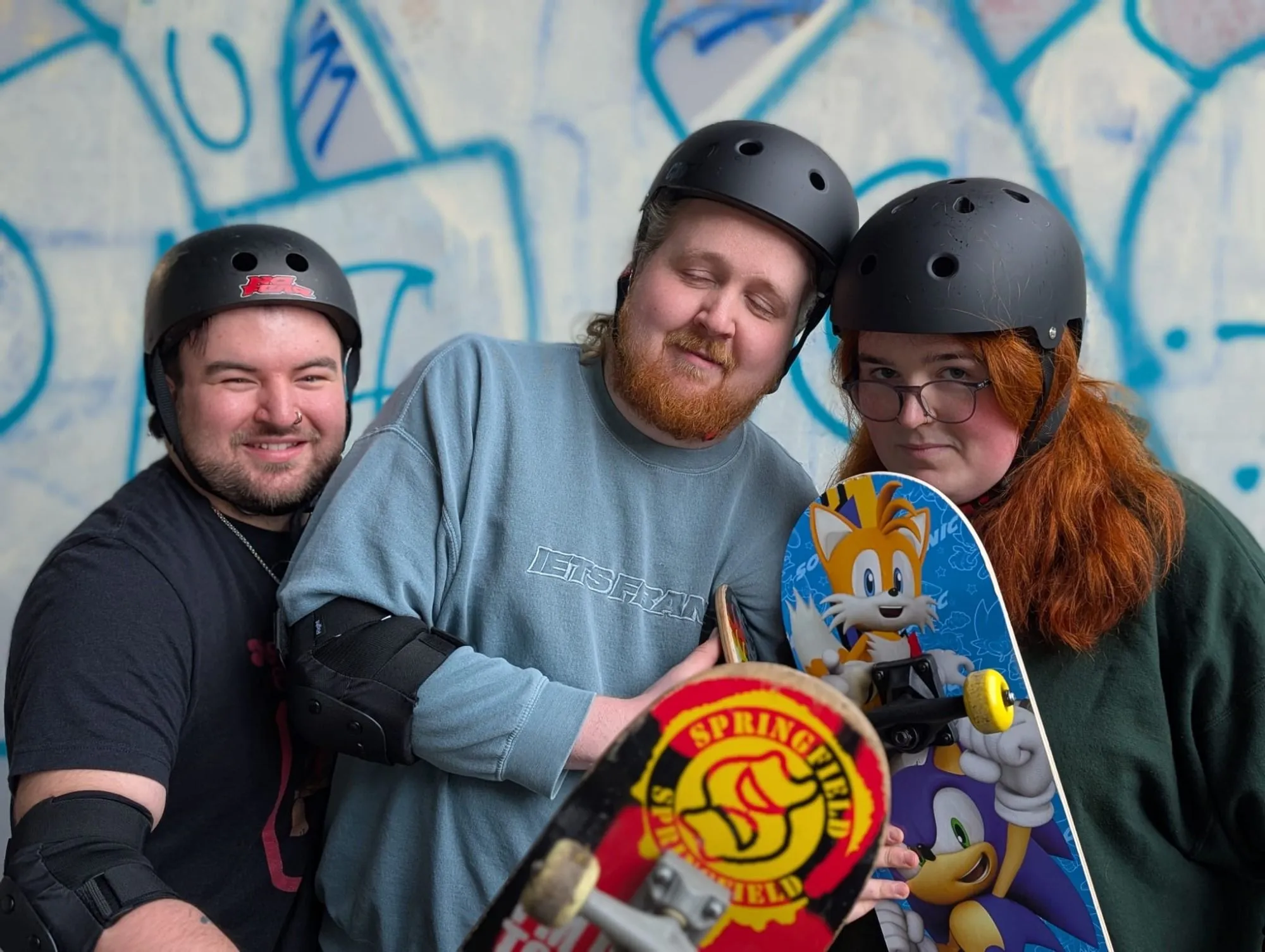 The three members of Pillow Fort wearing skate helmets in front of a graffiti wall, one holding a skateboard